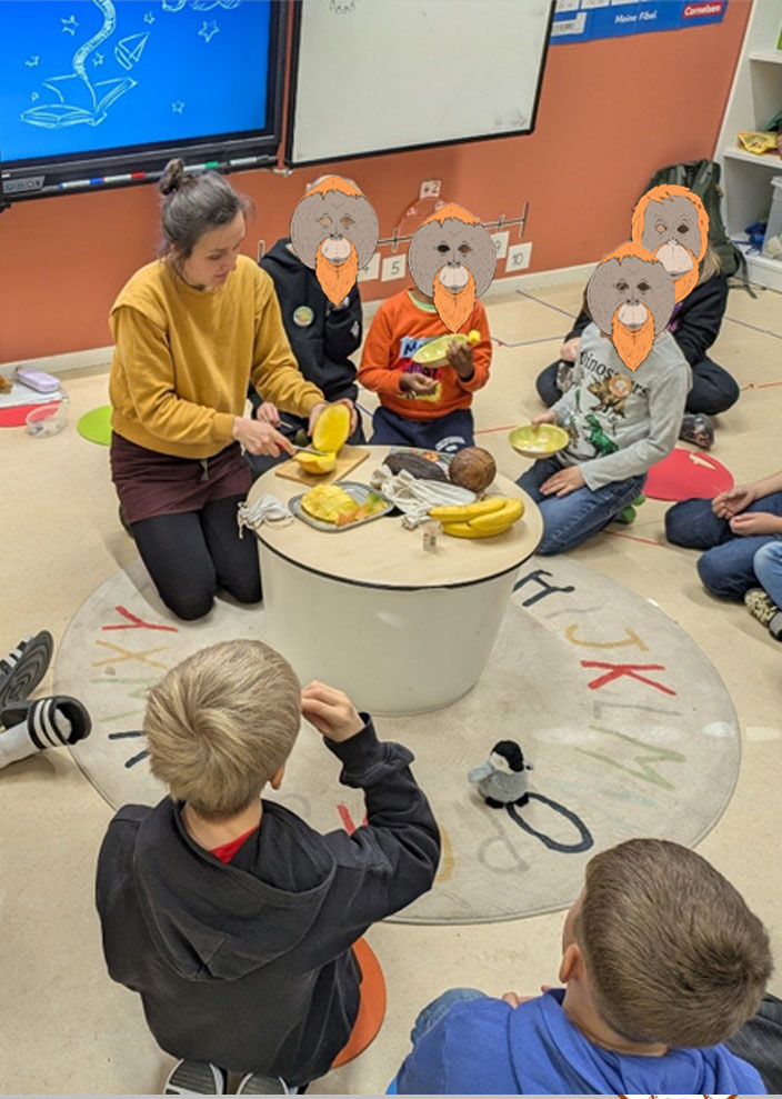 Mehrere Kinder sitzen im Kreis auf dem Boden. In der Mitte steht ein kleiner Tisch auf dem Bananen, eine Kokosnuss, eine Kakaofrucht, mehrere Stoffsäckchen und ein Schneidebrett liegen. Die Umweltbildnerin schneidet gerade Mango auf.