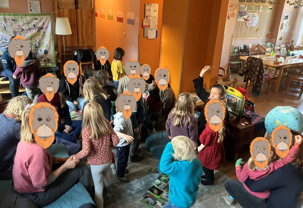 Mehrere Kinder stehen in einer Art Wohnzimmer. Um sie herum befinden sich einige Erwachsene. Am rechten Bildrand liegt ein Globus. Die Umweltbildnerin sitzt vor einem Bildertheater.