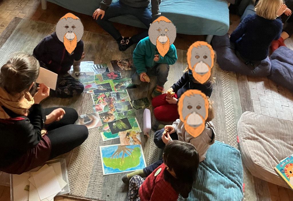 Mehrere Kinder stehen in einer Art Wohnzimmer. Um sie herum befinden sich einige Erwachsene. Am rechten Bildrand liegt ein Globus. Die Umweltbildnerin sitzt vor einem Bildertheater.