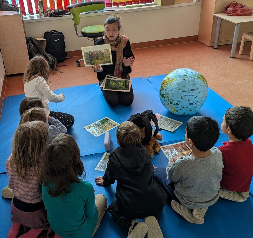 Kinder sitzen auf einer blauen Matte. Darauf liegen Bildkarten von Jahreszeiten, ein Rucksack und ein Globus. Die Umweltbildnerin hält ein Bild vom Regenwald nach oben.