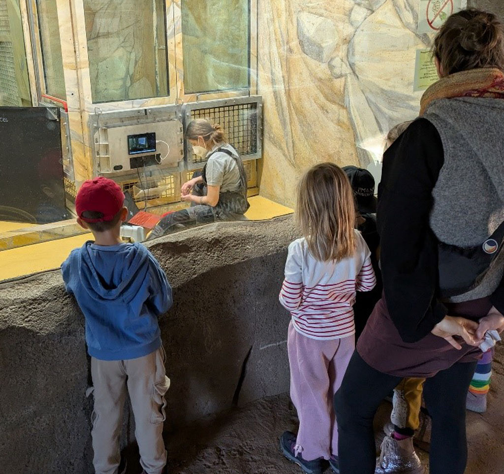 Mehrere Kinder und die Umweltbildnerin stehen vor einer Forschungsstation. Hinter der Schutzscheibe befinden sich Käfige, eine Forscherin, ein Schimpanse und ein Bildschirm.