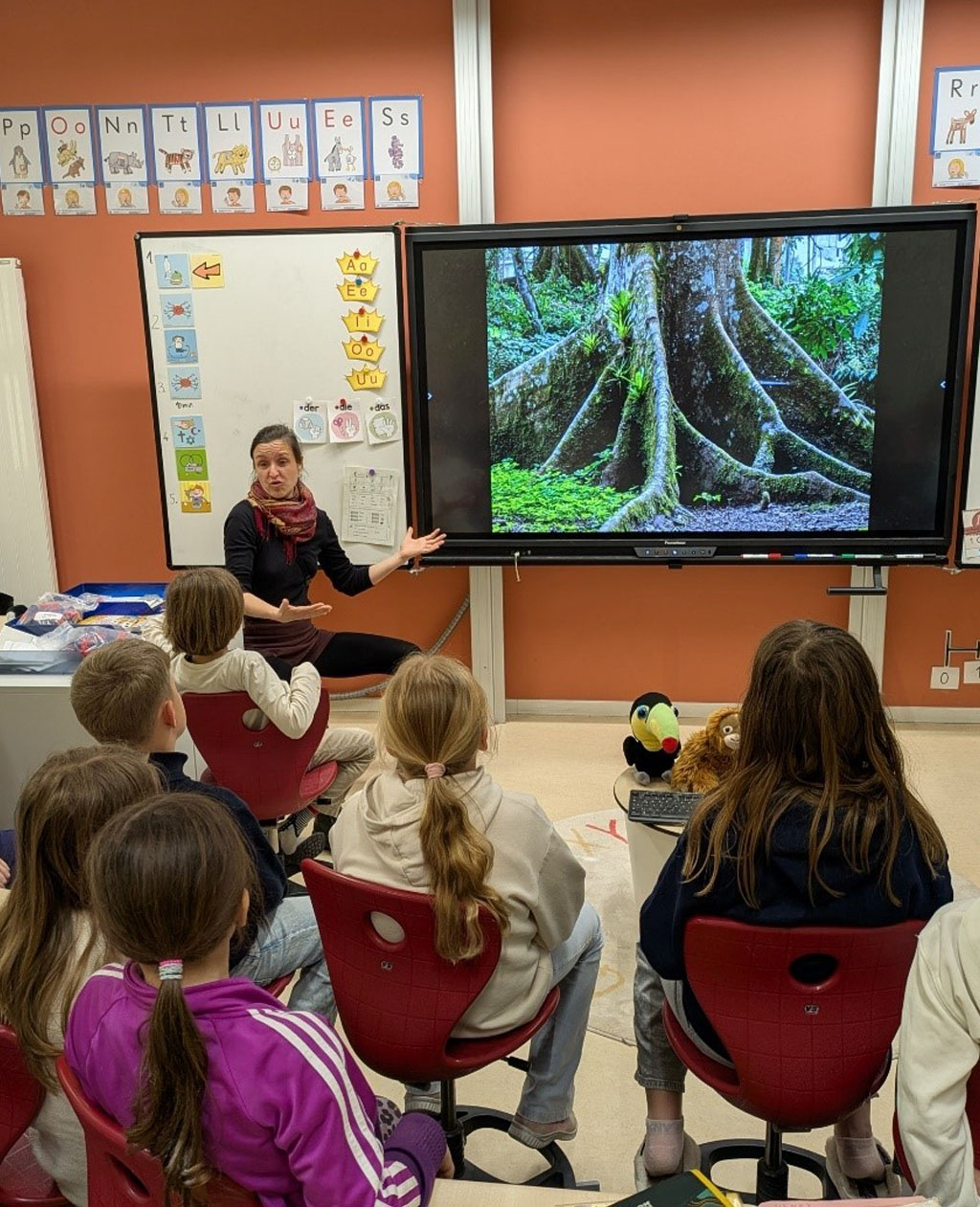 Mehrere Kinder sitzen in Reihen und schauen zu einer digitalen Tafel. Auf dieser wird gerade das Bild eines Brettwurzlers gezeigt. Die Umweltbildnerin sitzt daneben und erklärt das Gezeigte.