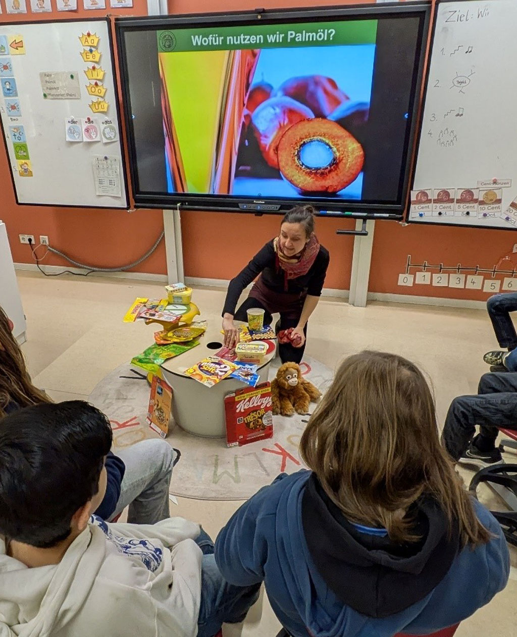 Mehrere Kinder sitzen im Halbkreis um die digitale Tafel. Auf dieser sind Palmölfrüchte und Öl abgebildet und es ist die Frage angeschrieben: Wofür nutzen wir Palmöl? Die Umweltbildnerin sitzt an einem zentralen Tisch und zeigt verschiedene Produktverpackungen.