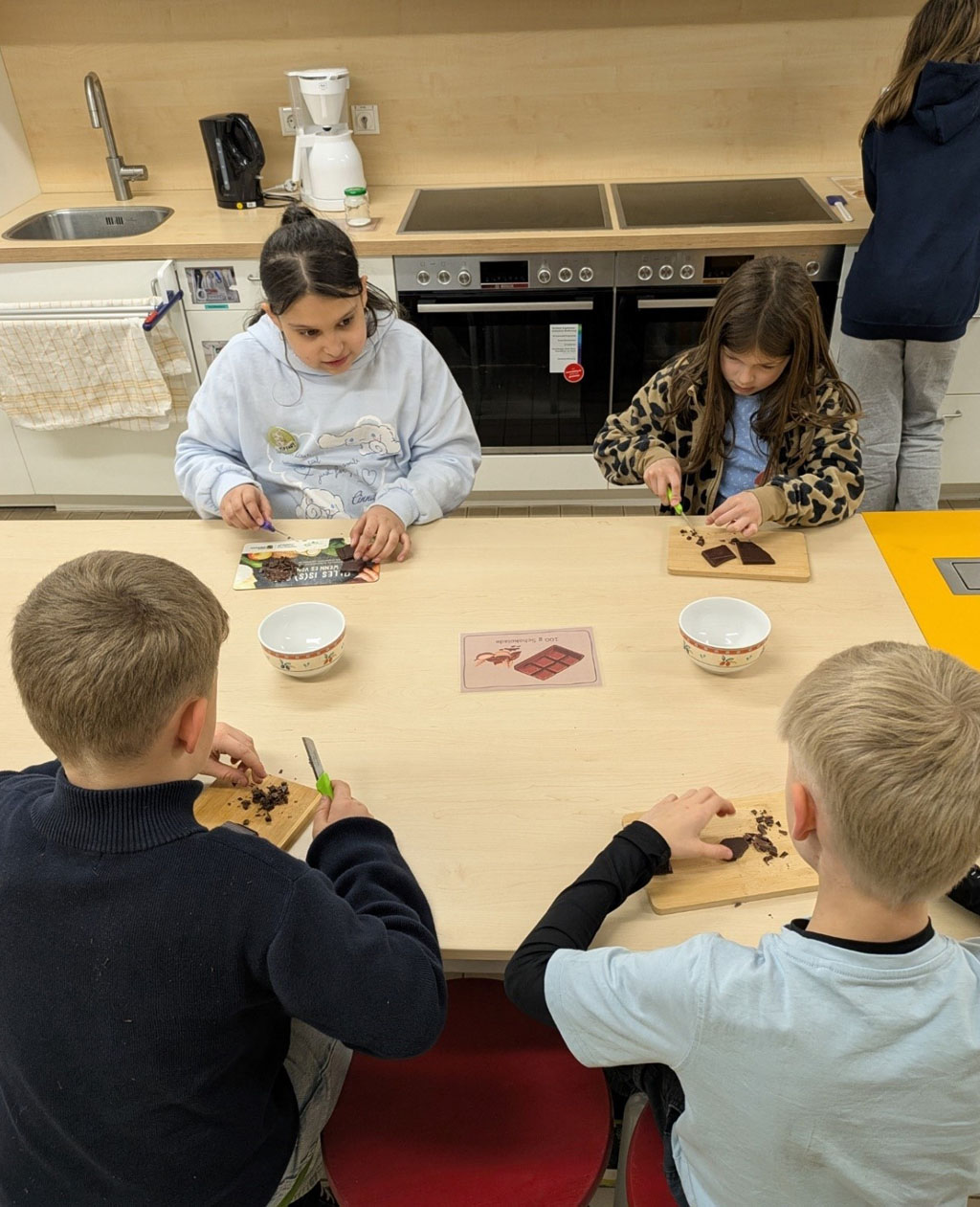 Vier Kinder sitzen in einer Küche und zerkleinern Schokolade. In der Mitte des Tisches liegt eine Bildkarte. Im Hintergrund steht eine weitere Person.