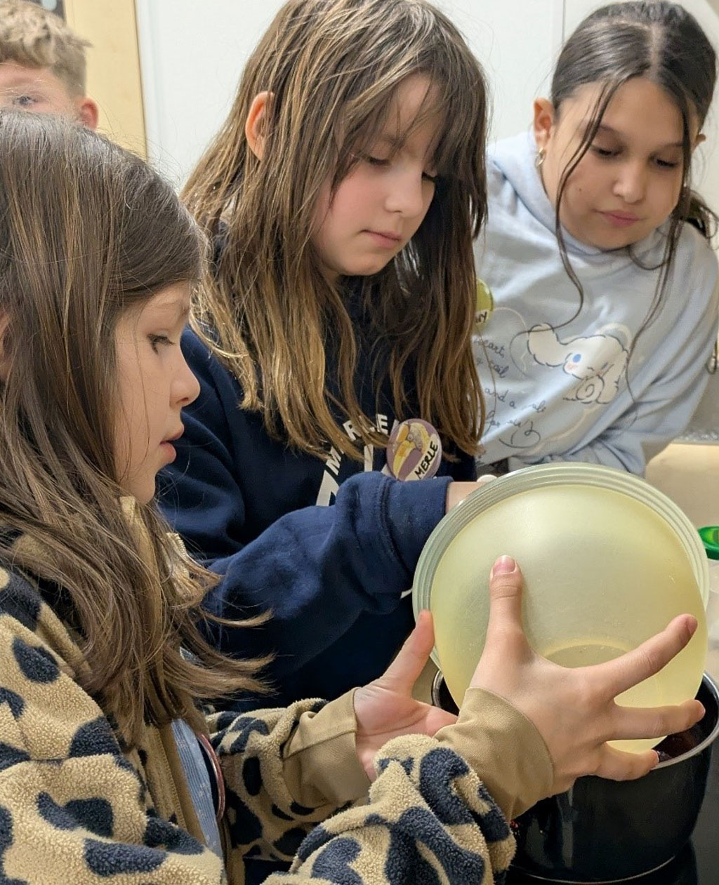 Drei Kinder stehen an einem Herd. Eines der Kinder füllt etwas aus einer Schüssel in einen Topf.
