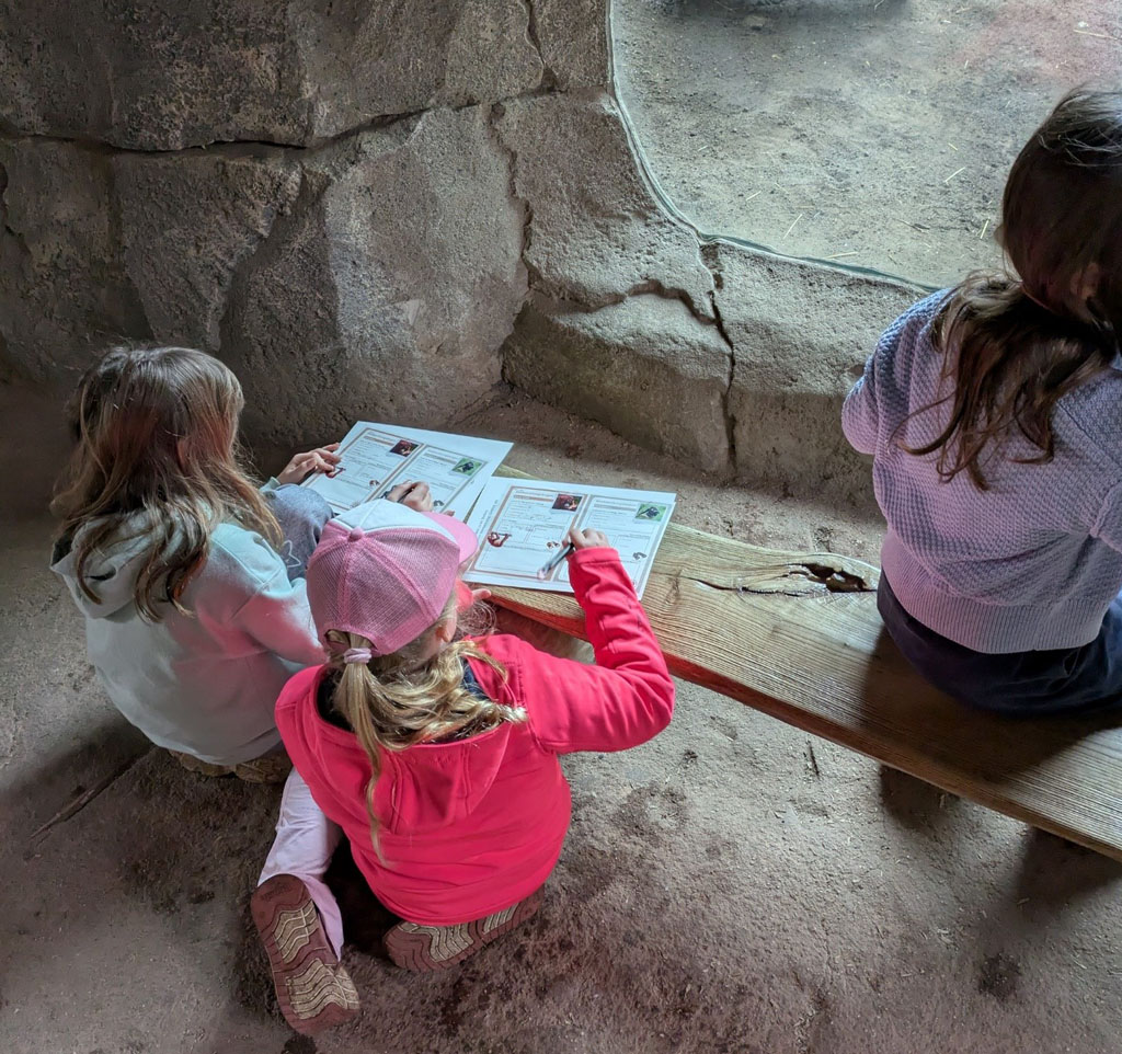 Zwei Kinder hocken vor einer Bank und füllen einen Beobachtungsbogen aus. Auf der Bank sitzt eine weitere Person.