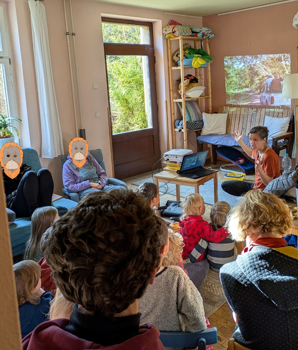 Im Familienzentrum Plagwitz sitzen mehrere Familien im offenen Wohnzimmer. Die meisten Kinder sitzen auf dem Boden. Über einem Sofa befindet sich eine Projektion. Die Umweltbildnerin hockt davor und erklärt etwas.