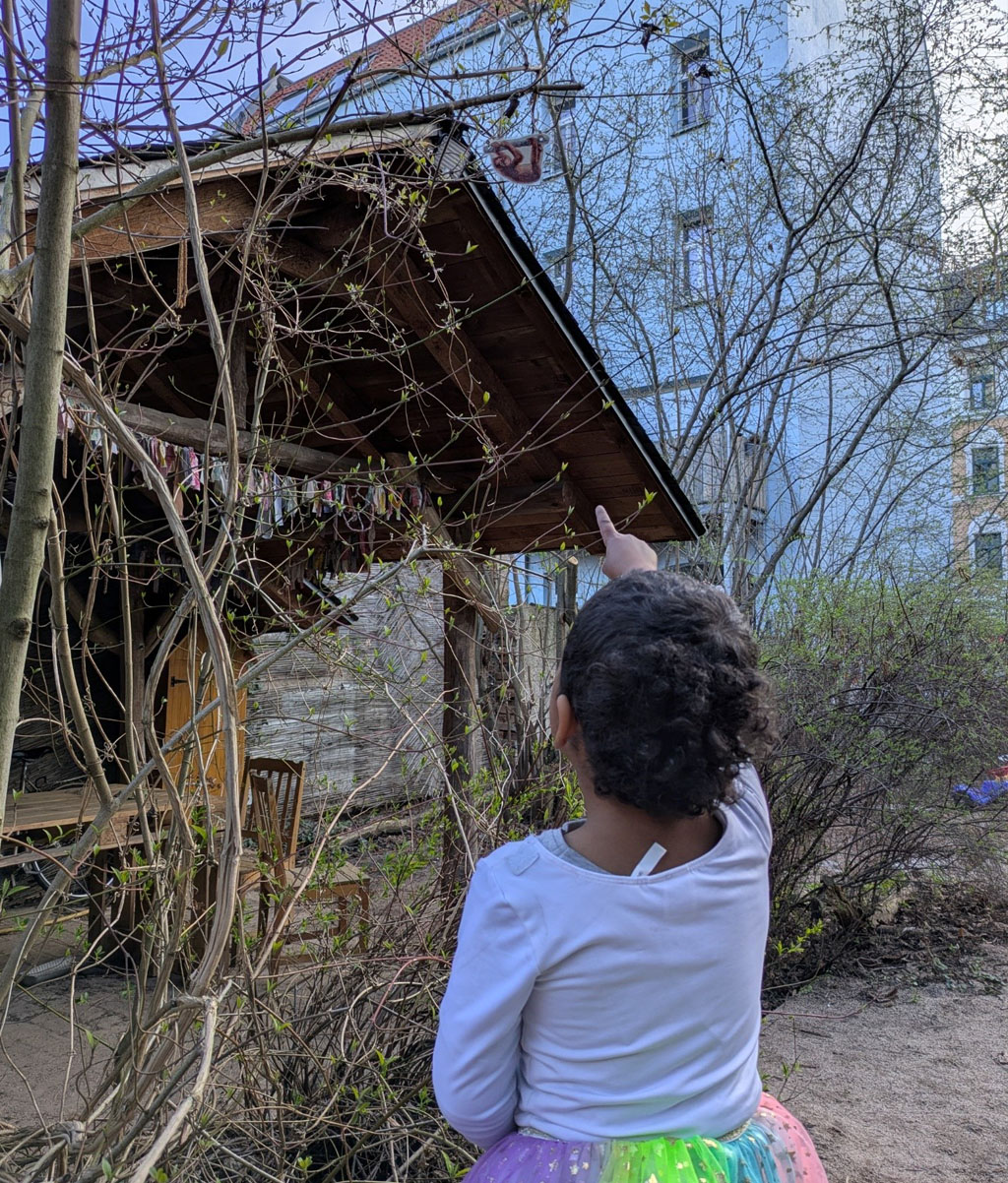 Ein Kind steht in einem Garten. Im Hintergrund befindet sich eine Holzhütte. Das Kind zeigt auf ein Orang-Utan-Bild, welches in einem Baum über ihm hängt.