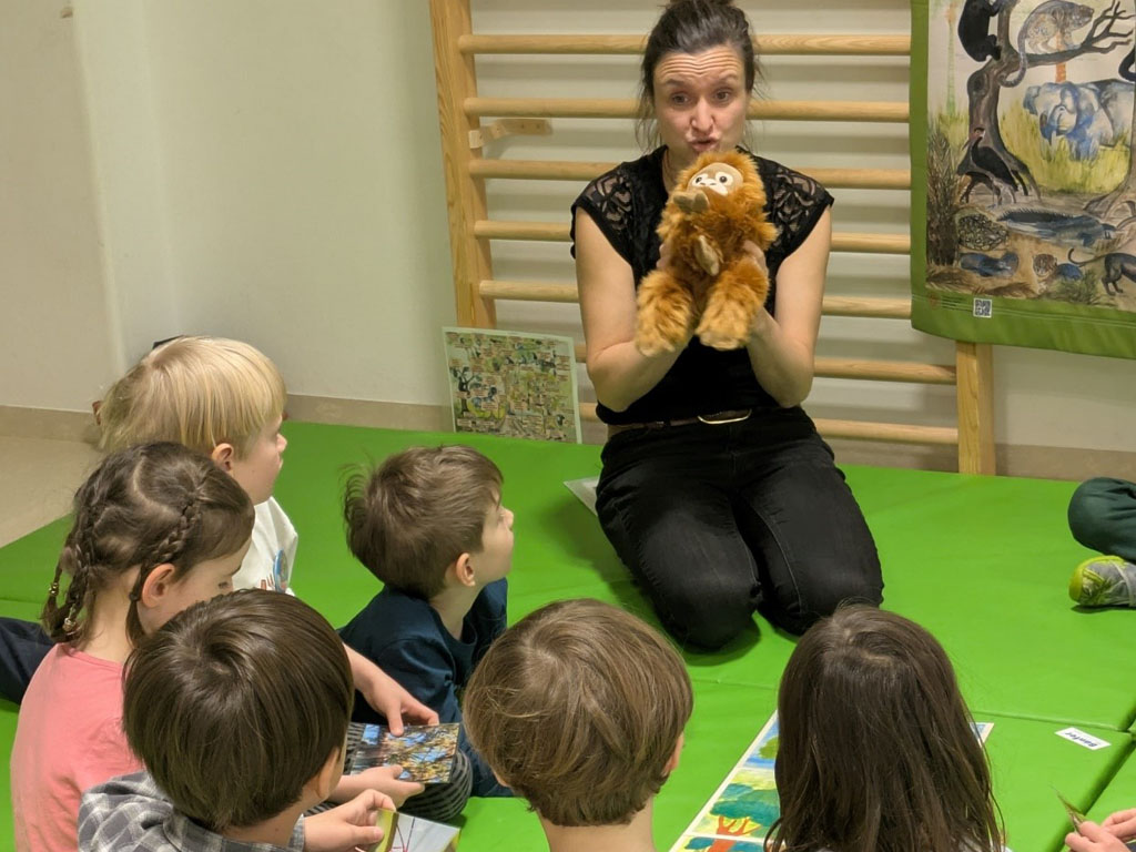 Kinder sitzen in einem Kreis auf grünen Matten. Die Umweltbildnerin hält ein Orang-Utan-Kuscheltier hoch. Dahinter hängt ein Plakat mit Tierabbildungen.