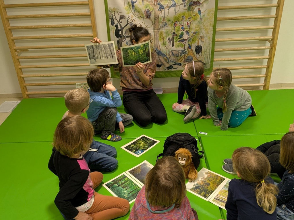 Kinder sitzen in einem Kreis auf grünen Matten. In der Mitte liegt ein schwarzer Rucksack, ein Orang-Utan-Plüschtier und Bildkarten. Die Umweltbildnerin hält zwei Bildkarten hoch. Auf der einen ist ein verschneiter Winterwald abgebildet, auf der anderen Regenwald. Dahinter hängt ein Plakat mit Tierabbildungen.