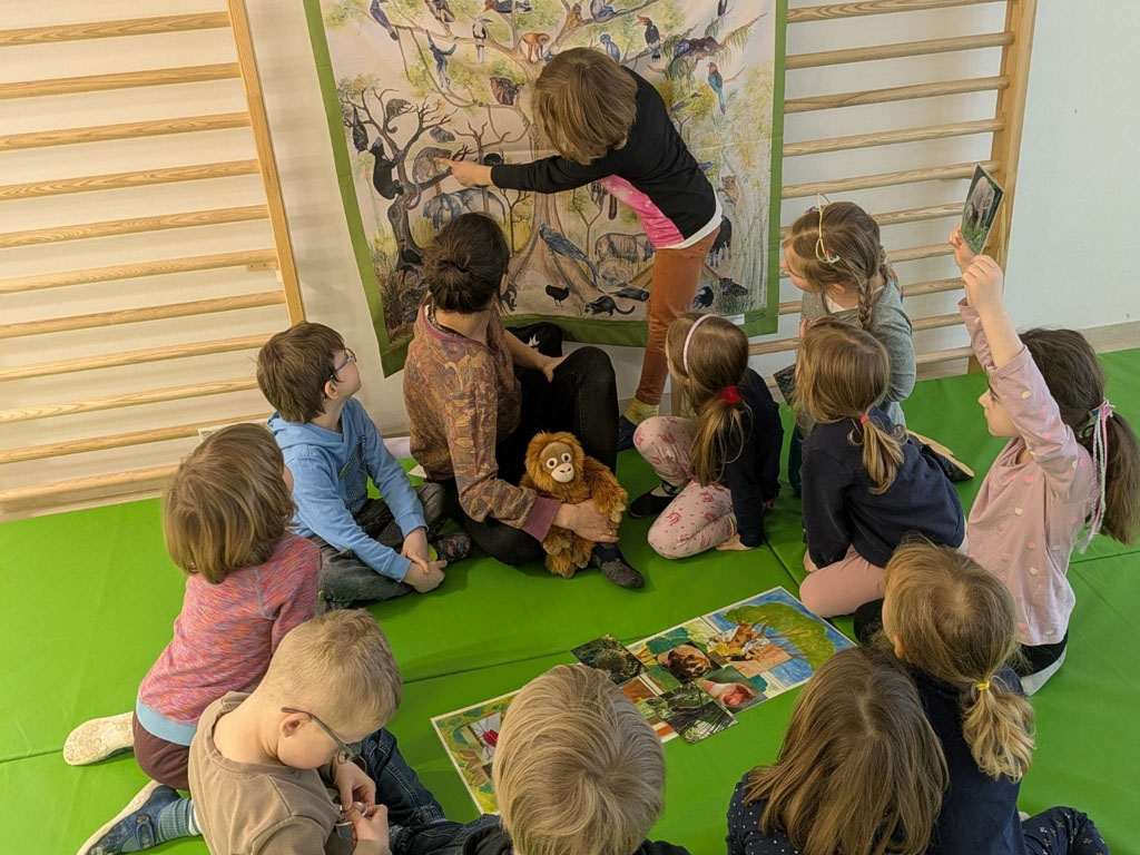 Kinder sitzen in einem Kreis auf grünen Matten. Die Umweltbildnerin hält ein Orang-Utan-Kuscheltier. Dahinter hängt ein Plakat mit Tierabbildungen. Ein Kind zeigt auf eines der Tiere auf dem Plakat.