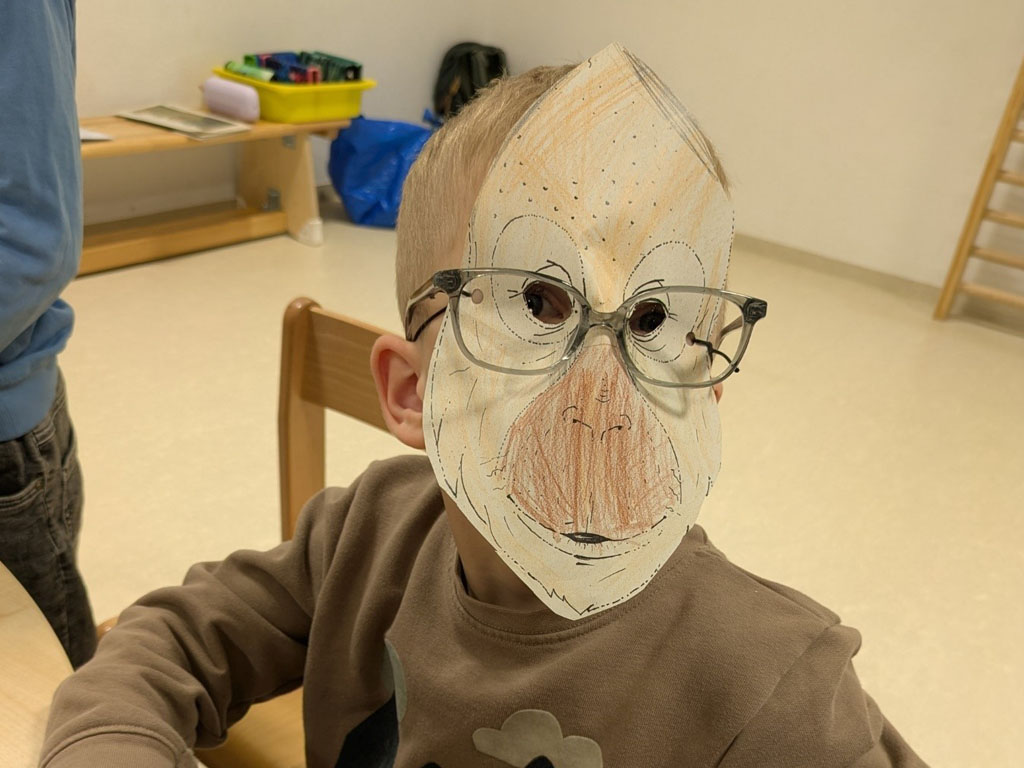 Ein Kind sitzt auf einem Stuhl und trägt über seiner ausgemalten Orang-Utan-Maske seine Brille.