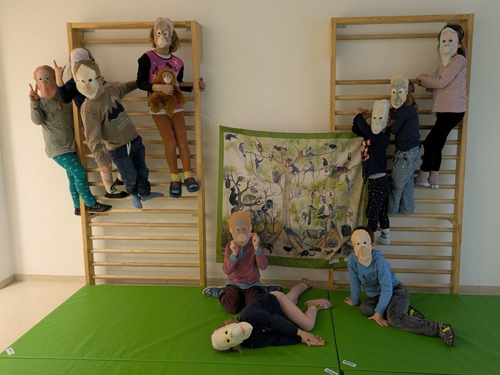 Kinder stehen auf zwei Sprossenwänden. Dazwischen hängt ein Plakat mit Tieren und Bäumen. Davor liegen drei Kinder auf grünen Sportmatten. Alle Kinder tragen Orang-Utan-Masken.