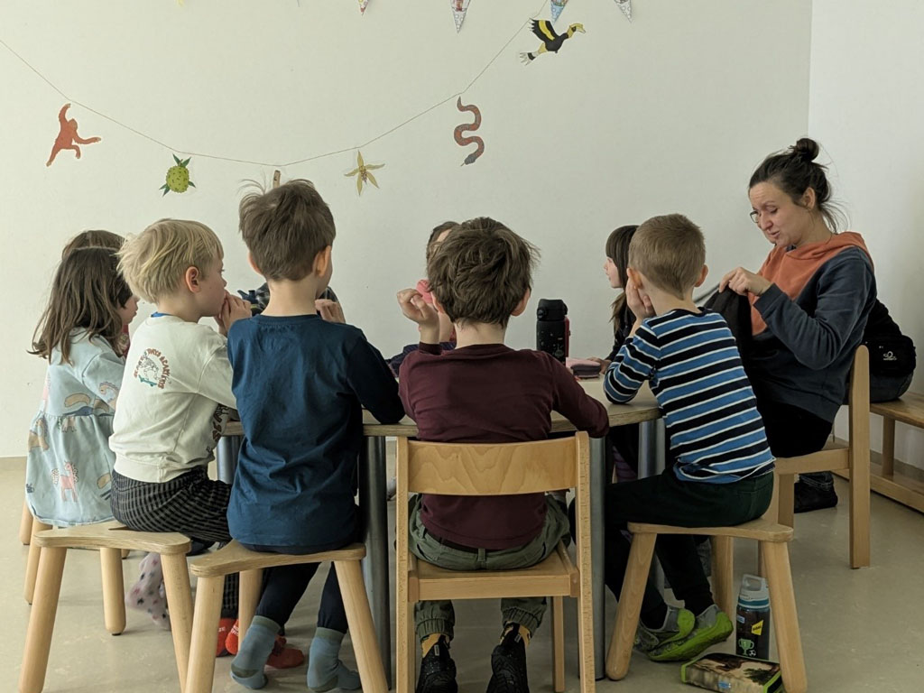 Kinder sitzen an einem Tisch. Darüber hängt eine Girlande mit Früchten, Tieren und Blüten des Regenwaldes.