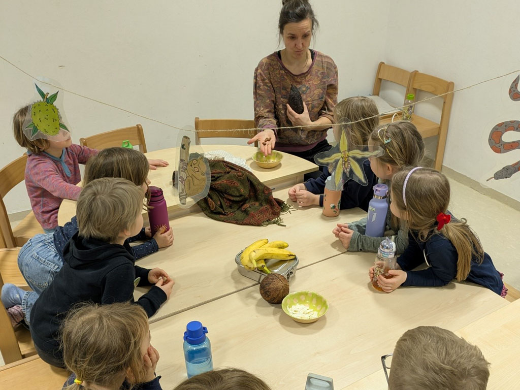 Kinder sitzen um einen Tisch. Darauf liegen Bananen, eine Kokosnuss, eine Schale mit Kokosnusschips und verschiedene Trinkflaschen. Die Umweltbildnerin hält in der einen Hand eine Kakaofrucht und zeigt auf ihrer anderen einzelne Kakaobohnen.