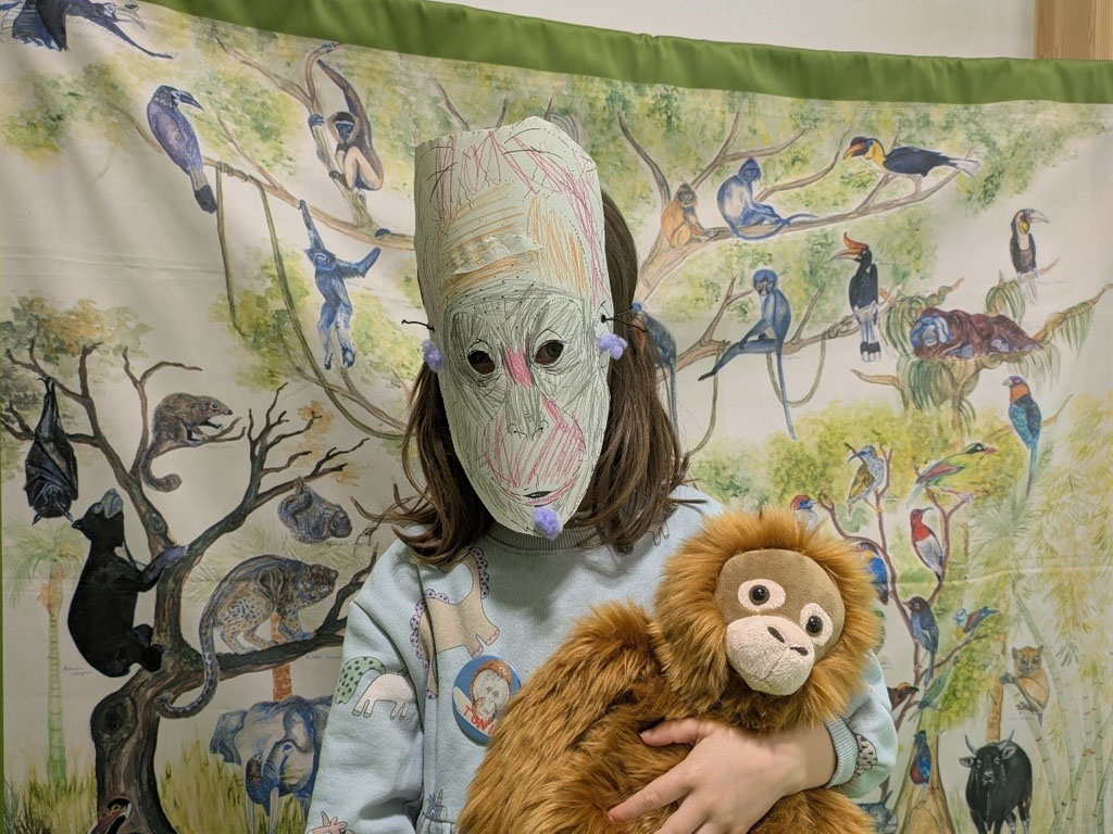 Ein Kind steht vor einem Regenwaldplakat und hält einen Plüsch-Orang-Utan im Arm. Im Gesicht trägt es eine bunte Orang-Utan-Maske.