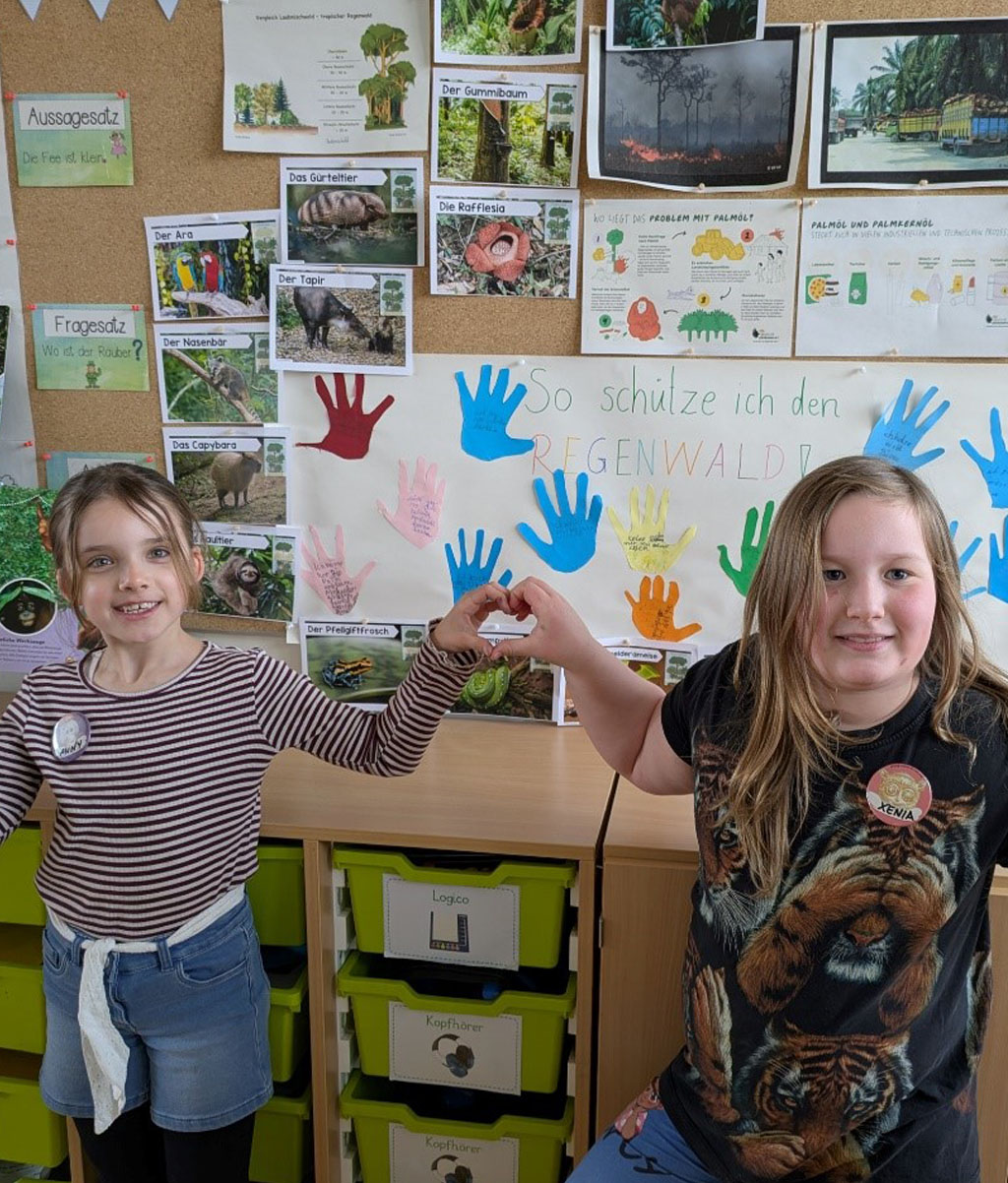 Zwei Kinder formen die Hände zu einem Herz. Sie stehen vor einer Pinwand, welche mit einem Plakat zum Thema: „So schütze ich den Regenwald“ und vielen Tier- und Pflanzenbildern behangen ist.