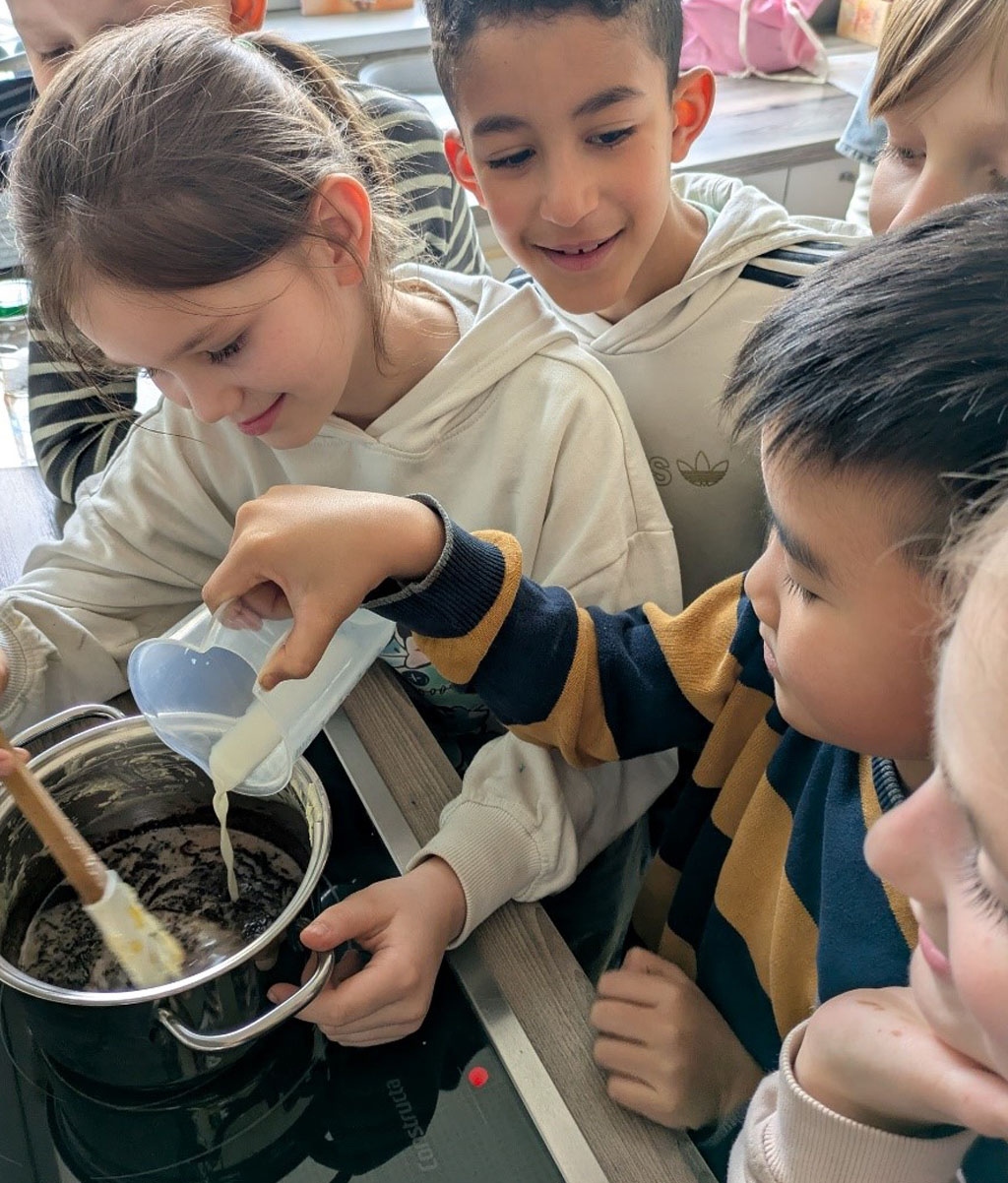 Sechs Kinder stehen an einem Herd. Ein Kind gießt Milch in einen Topf. Ein anderes Kind rührt die Schokoladencreme um.