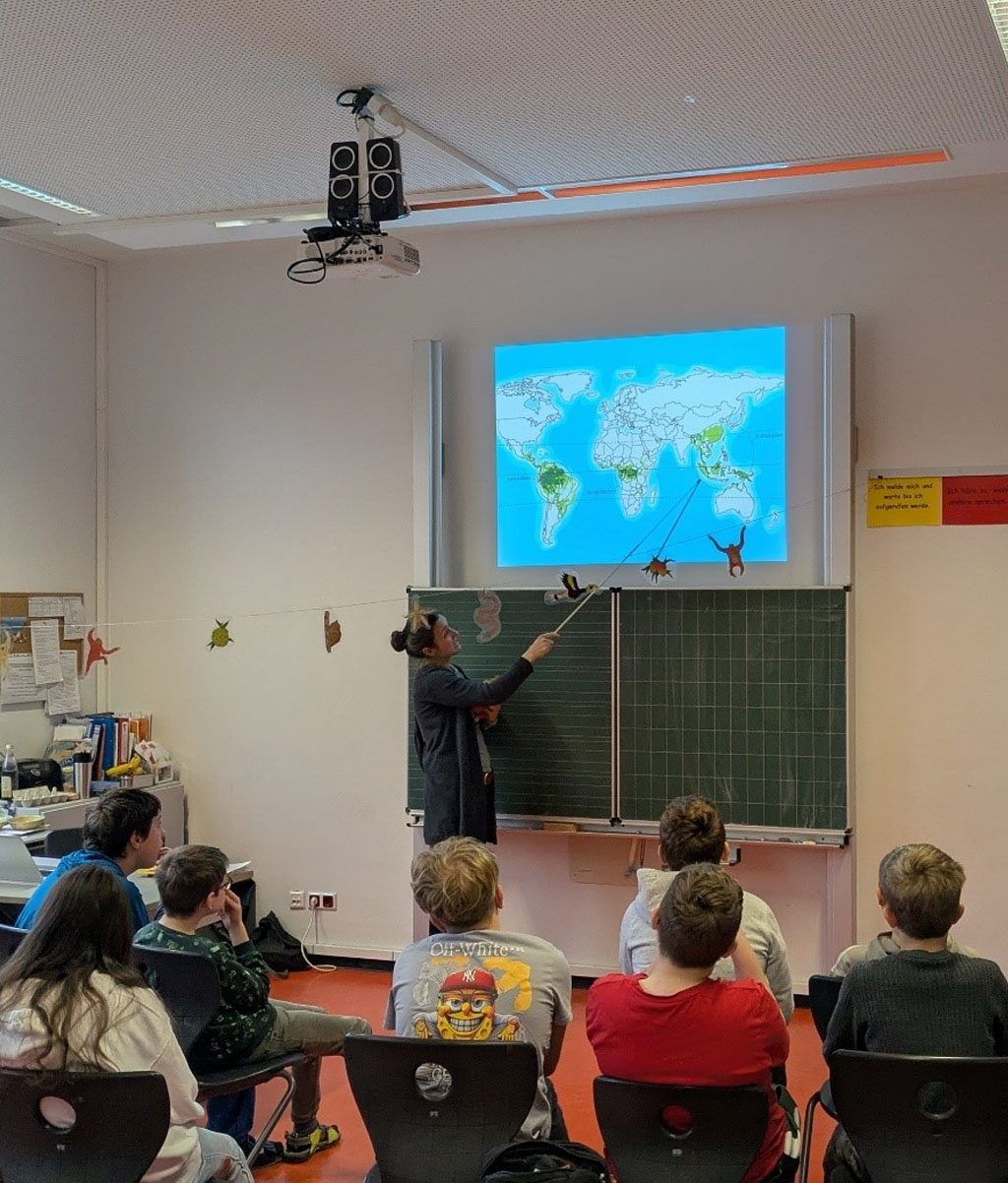 In einem Klassenzimmer befinden sich mehrere Kinder. Über der Tafel wird eine Präsentation projiziert. Die Umweltbildnerin zeigt auf einer Weltkarte das südostasiatische Regenwaldgebiet.