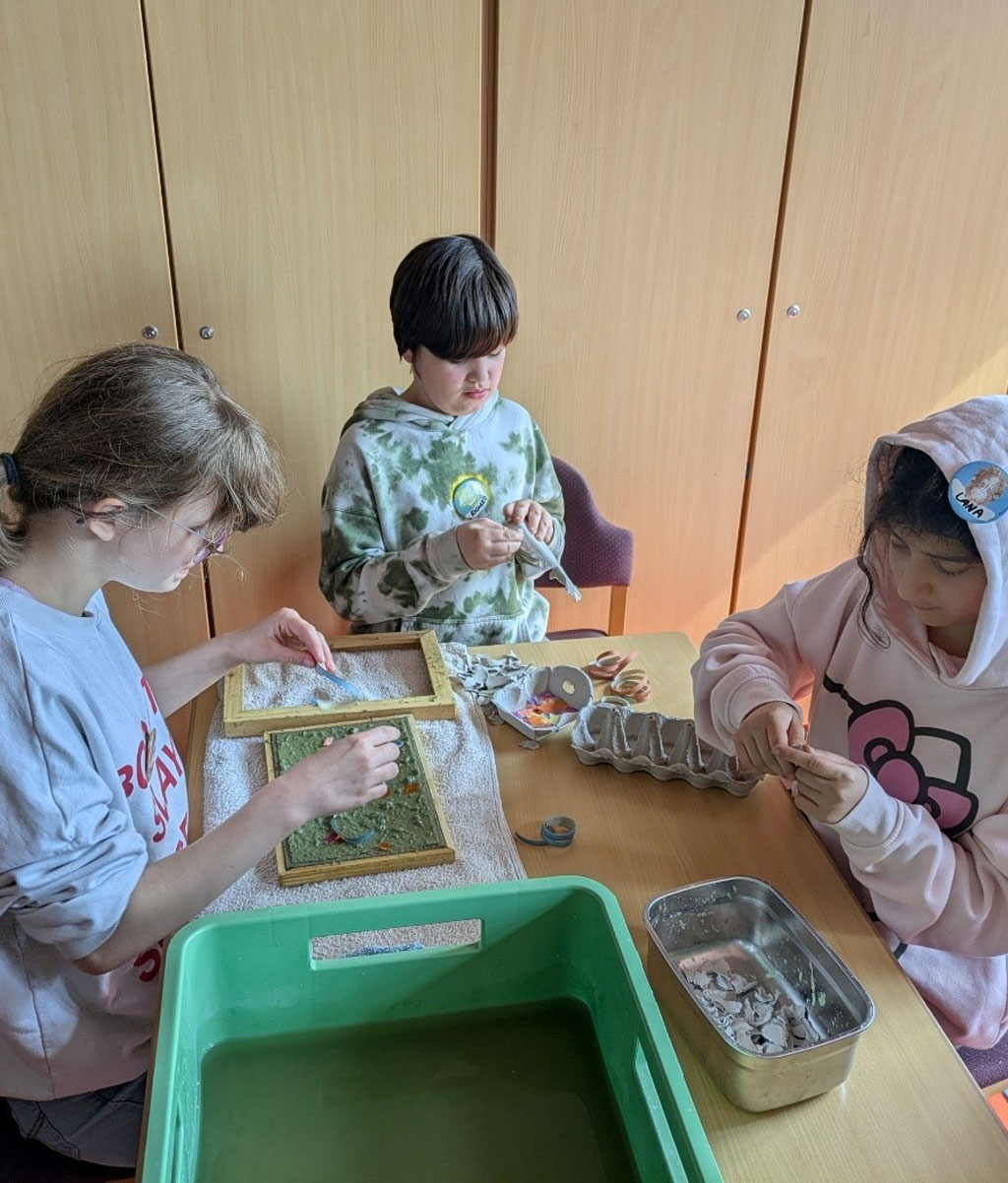 Drei Kinder sitzen an einem Tisch. Darauf stehen eine große grüne Wanne mit Pulpe darin, Eierpappen, Luftschlangen und ein Handtuch, auf dem ein Papierschöpfrahmen liegt. Zwei Kinder reißen die Eierpappen in kleine Stücke. Ein Kind gestaltet ein frisch geschöpftes Papier mit Konfetti.