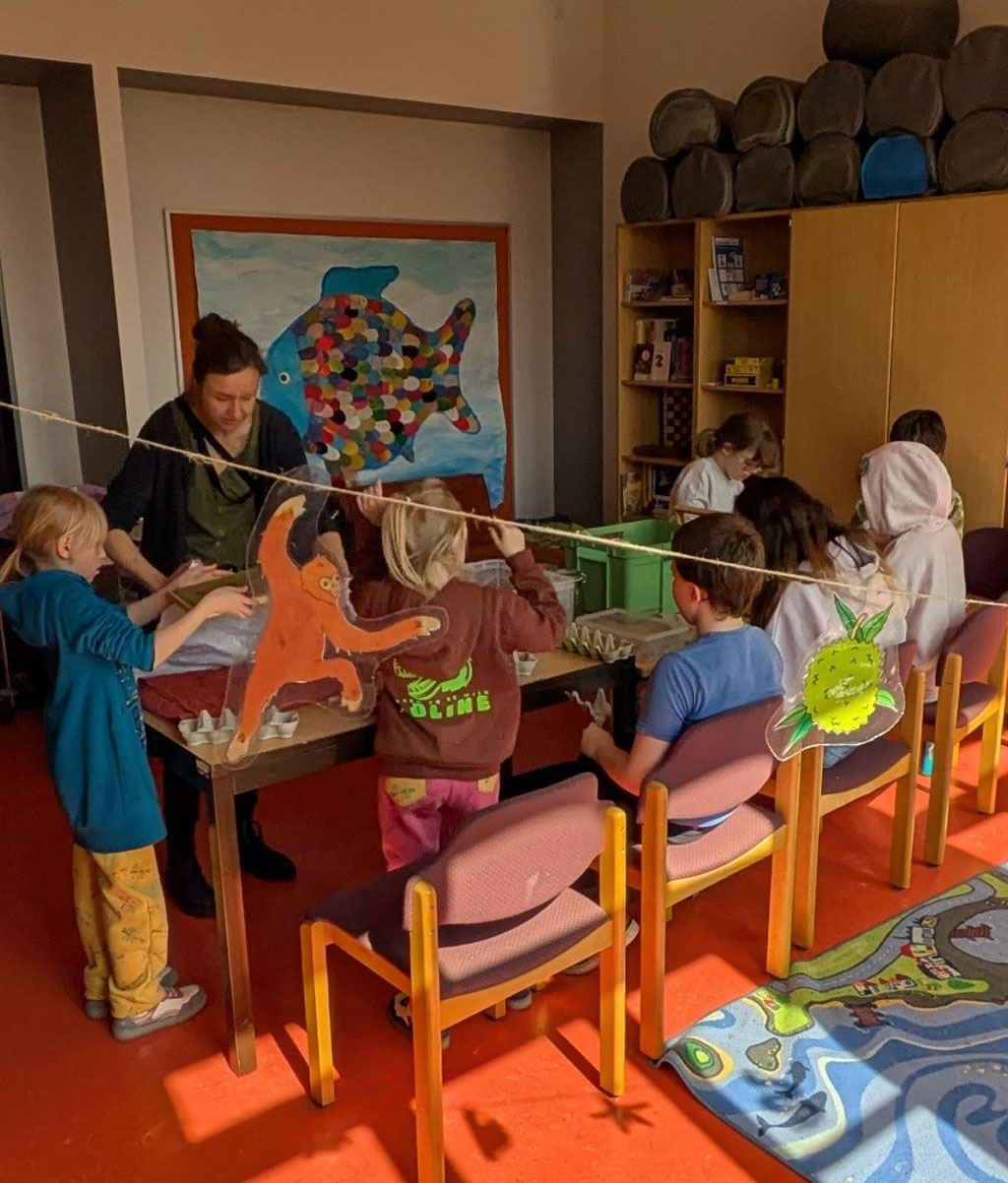 Kinder sitzen um einen langen Tisch herum. Die Umweltbildnerin steht neben einem Kind, das einen Papierschöpfrahmen in die Luft hält. Im Vordergrund hängt eine Girlande mit Regenwaldtieren und -früchten.