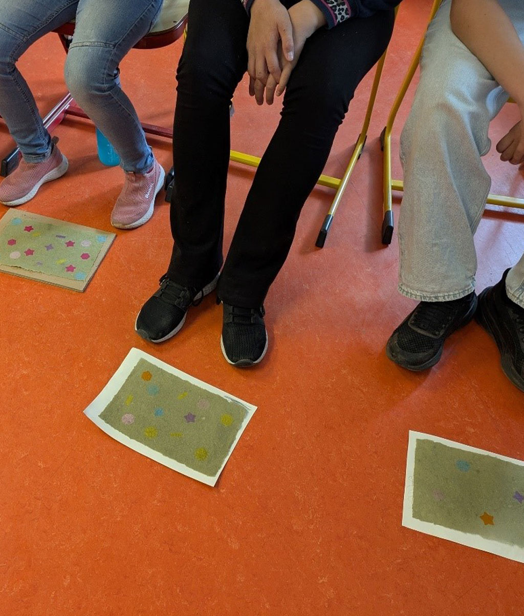 Vier Kinder sitzen auf Stühlen. Vor ihnen liegen frisch geschöpfte Papiere auf dem roten Fußboden.