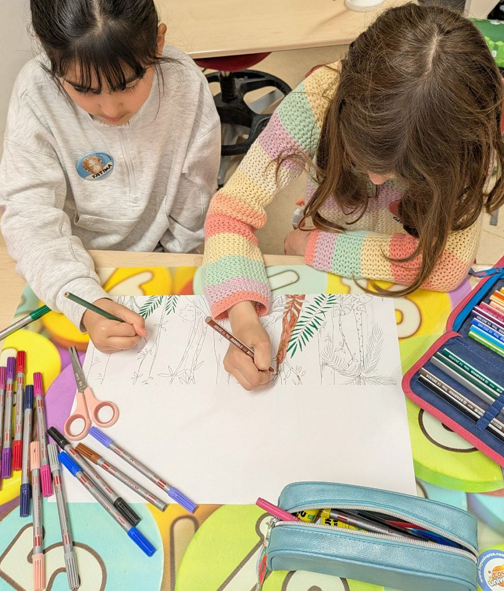 Zwei Kinder sitzen an einem Tisch und malen gemeinsam ein Regenwaldbild aus. Um das Bild herum liegen Stifte und Federmappen sowie eine Schere.