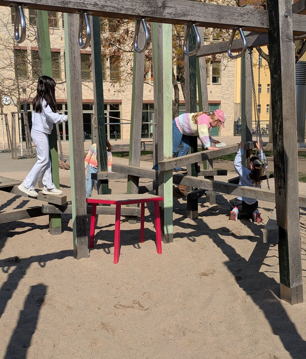 Auf einem Klettergerüst hangeln, balancieren und bewegen sich drei Kinder. Eines der Kinder hat eine Karte gefunden und liest gerade, was darauf steht. Im Hintergrund läuft ein weiteres Kind.