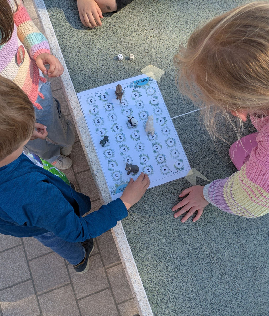 Auf einer Tischtennisplatte klebt ein einfaches Spielfeld mit den Felden eins bis 40. Auf dem Spielfeld befinden sich mehrere Tierfiguren auf unterschiedlichen Feldern. Daneben liegen zwei Würfel. Rings um das Spielfeld befinden sich mehrere Kinder.