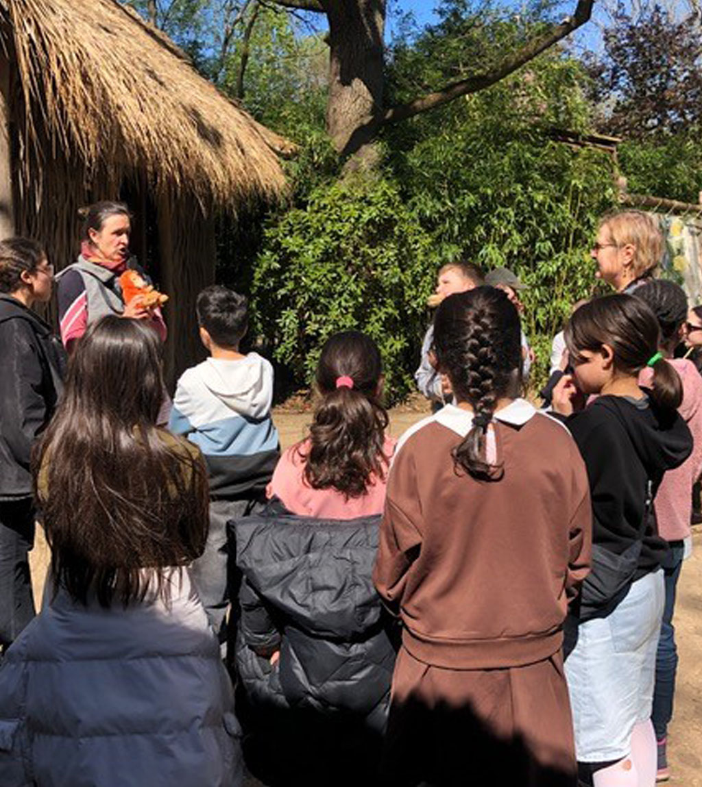 Die Umweltbildnerin hält einen Plüsch-Orang-Utan in der Hand. Um sie herum stehen im Halbkreis mehrere Kinder. Im Hintergrund steht eine Hütte mit Strohdach.