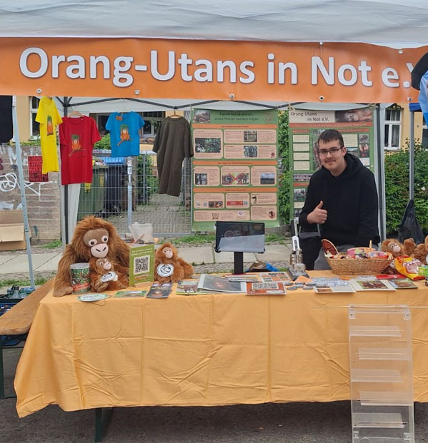 Zwischen zwei Ständen steht ein weißer Pavillon an dessen oberer Kante ein Banner mit der Aufschrift „Orang-Utans in Not e.V.“ hängt. Rechts davon steht eine Beachflag mit dem Vereinslogo und dem Foto eines Regenwaldes. Im vorderen Bereich steht ein Biertisch mit oranger Tischdecke. Darauf liegen Flyer, Plüschtiere, Spendendosen und weitere Verkaufsartikel. Hinter dem Tisch steht ein Mann, der den Daumen nach oben zeigt. An der Decke des Pavillons hängen T-Shirts und Beutel. Im Hintergrund sind Plakate befestigt.