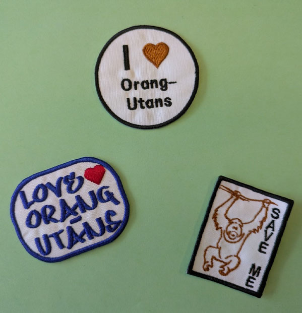 Auf grünem Untergrund liegen drei Aufnäher. Auf zweien davon steht „I love Orang-Utans“ auf dem dritten ist ein Orang-Utan schemenhaft abgebildet und daneben steht: „Save me“.