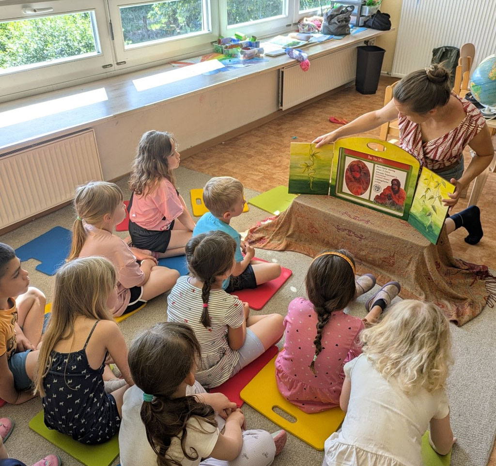 Kinder sitzen vor einem Kamishibai (Bildertheater) auf dem Boden. Die Umweltbildnerin klappt das Theater gerade auf.