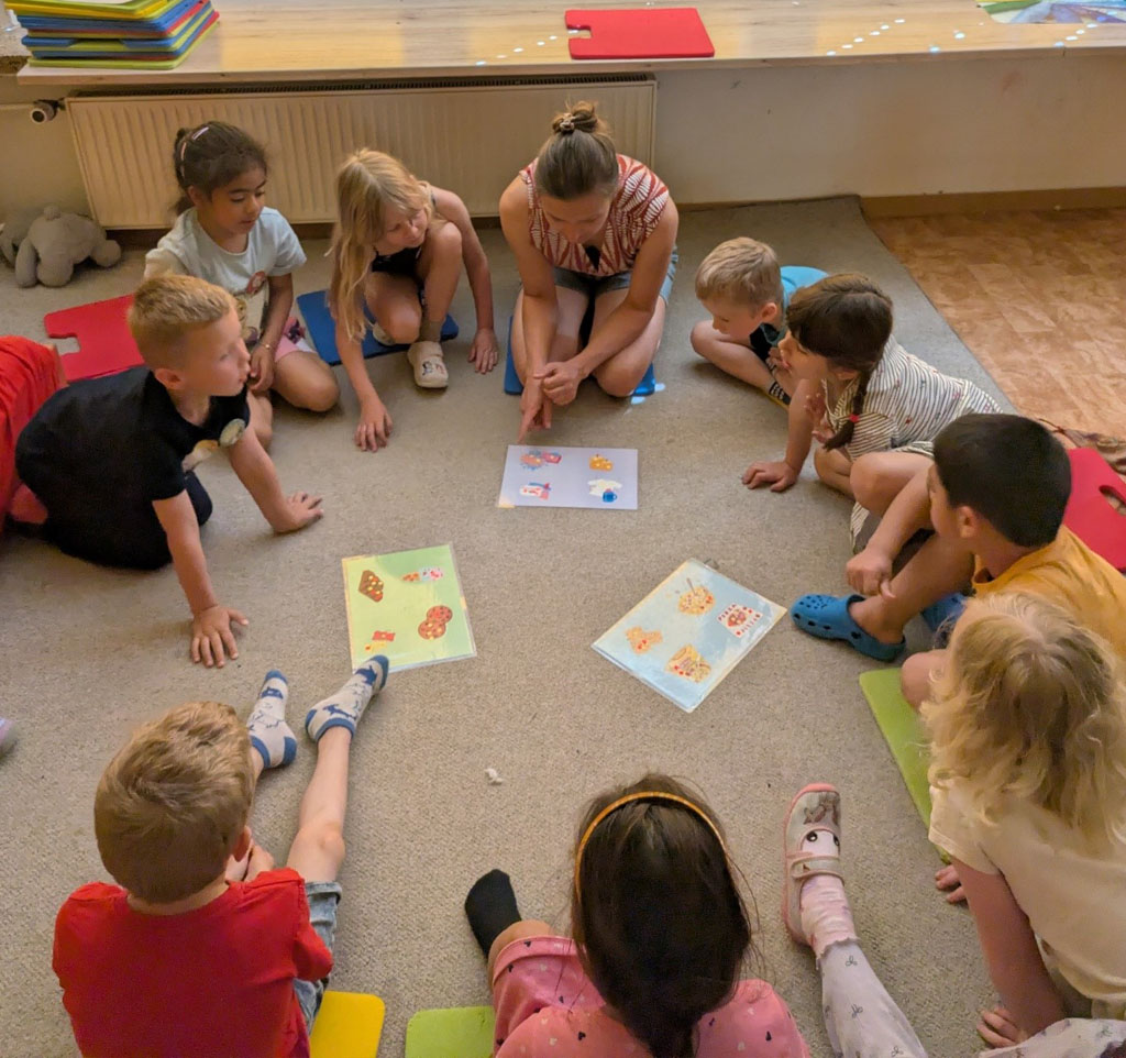 Kinder sitzen mit der Umweltbildnerin im Kreis auf dem Boden. In dessen Mitte drei Bildkarten liegen, auf denen verschiedene Produkte abgebildet sind.