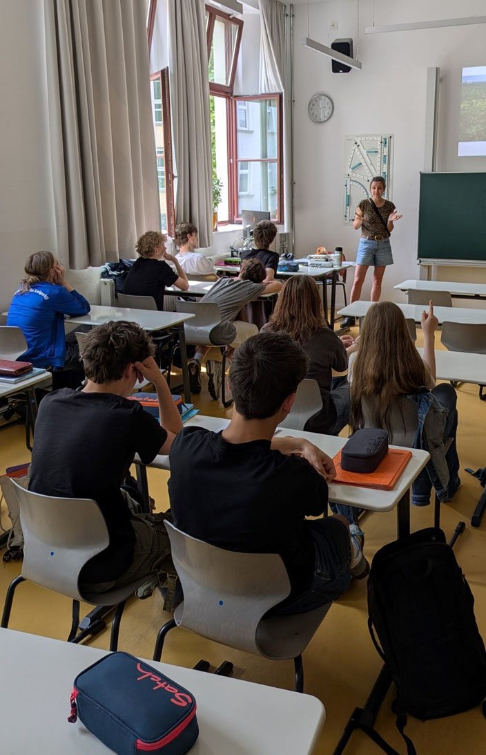 Jugendliche sitzen in einem Klassenzimmer. Die Umweltbildnerin steht vorn an der Tafel.