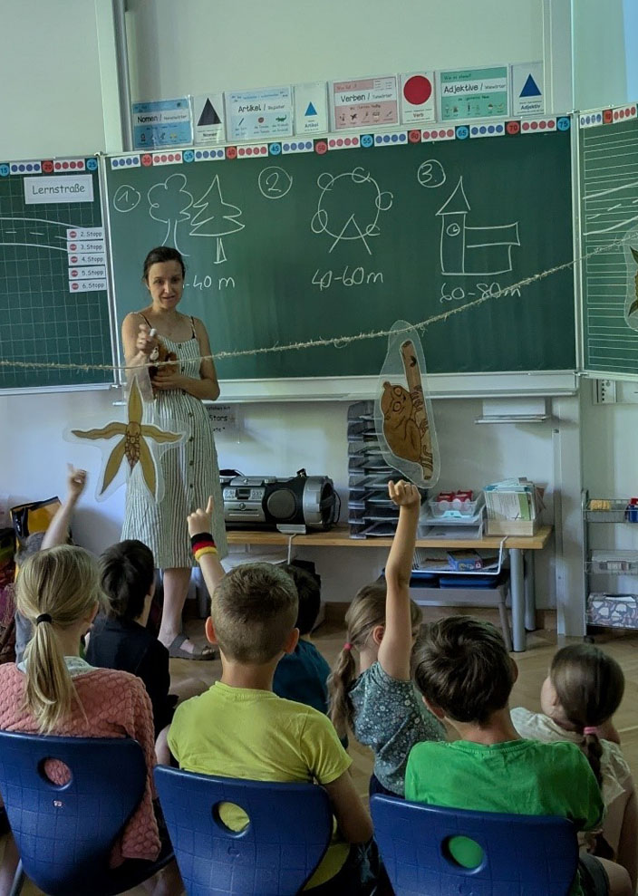 Kinder sitzen in einem Klassenzimmer und schauen in die Richtung Tafel. Vorn an der Tafel steht die Umweltbildnerin, einige Kinder melden sich.