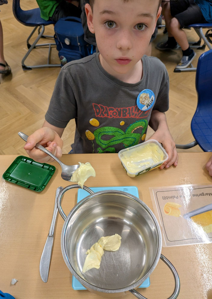 Ein Kind sitzt an einem Tisch und füllt Margarine in einen Topf. Auf dem Tisch liegt außerdem eine Bildkarte, ein Messer und eine Waage.