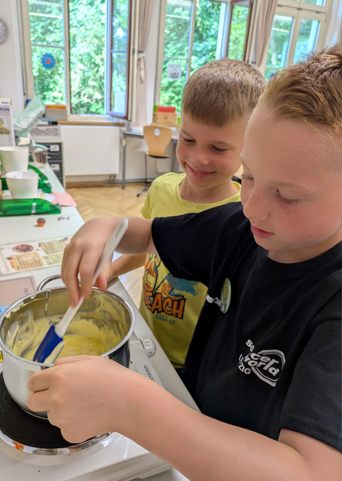 Zwei Kinder stehen an einer Herdplatte. Eines der Kinder rührt Margarine in einem Topf um. Das andere Kind sieht dabei zu.