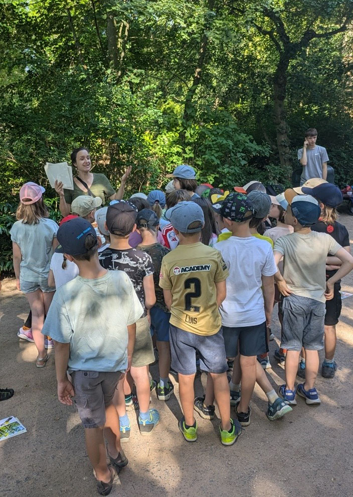 Eine Gruppe von ca. 30 Kindern steht als Traube auf einem Weg. Die Umweltbildnerin hält Dokumente in der Hand und erklärt gerade etwas.