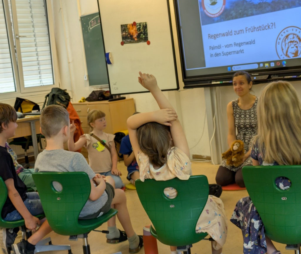 Kinder sitzen vor einer digitalen Tafel, auf der eine Präsentation läuft. Die Umweltbildnerin hockt vorn und hält ein Orang-Utan-Kuscheltier in den Händen. Zwei Kinder melden sich.