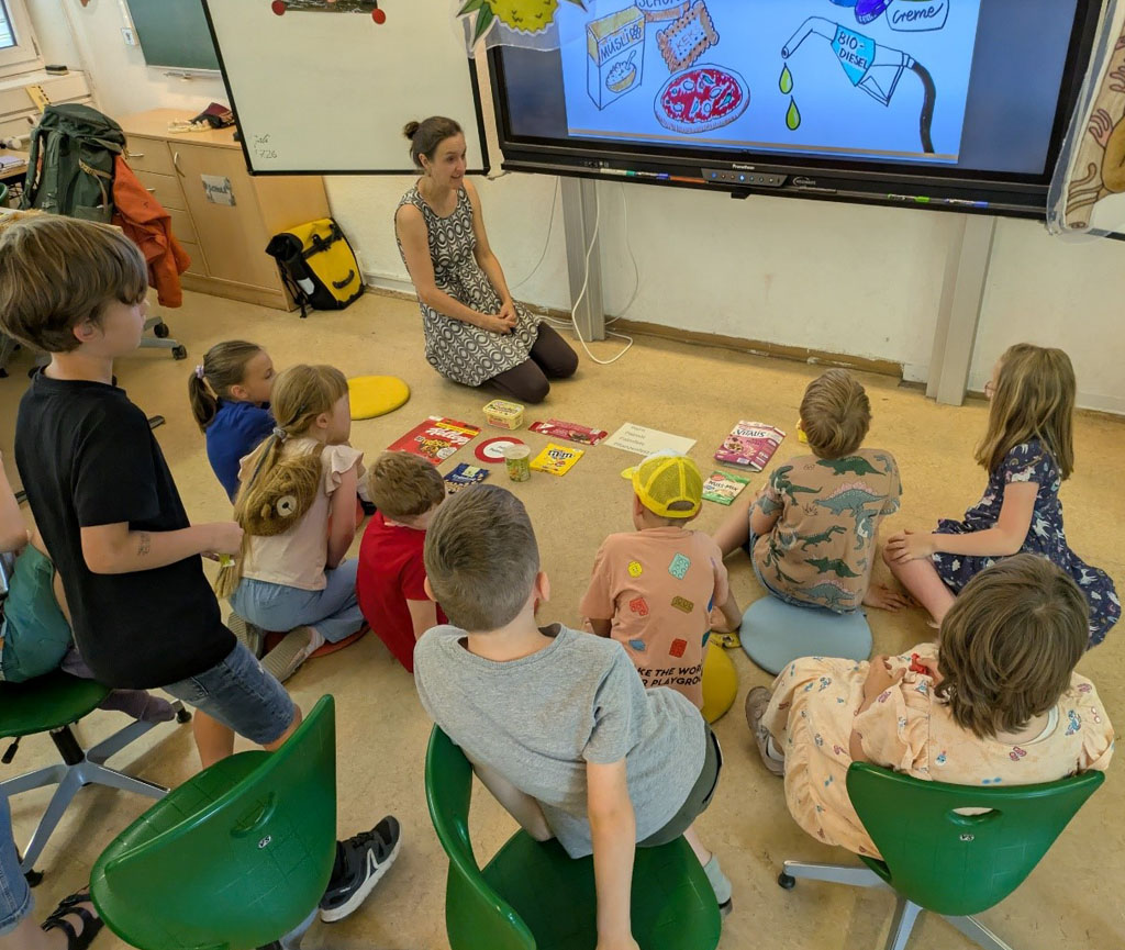 Kinder sitzen auf Stühlen und teilweise auf dem Boden vor einer digitalen Tafel, auf der eine Präsentation läuft. Die Umweltbildnerin sitzt vor Produktverpackungen, welche sortiert auf dem Boden liegen.