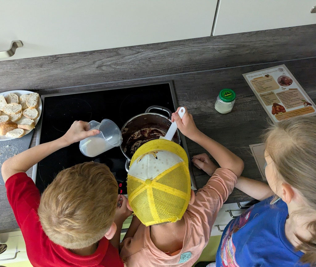 Drei Kinder stehen an einem Herd. Das linke Kind gießt Milch in einen Topf, das mittlere rührt darin um. Links vom Herd steht ein Teller mit Baguette-Scheiben, rechts liegt das Rezept für Palmölfreien Schokostreich.