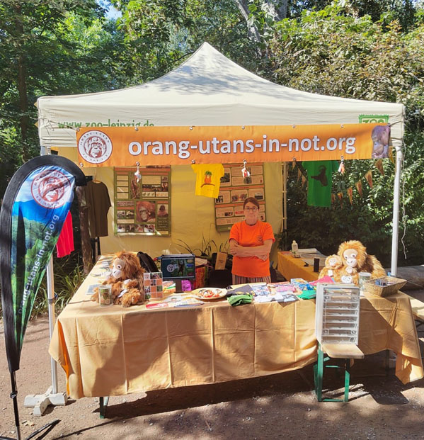 Unter einem Pavillon des Orang-Utan-in Not e.V. stehen drei Tische mit Plüschtieren, T-shirts, Bastel- und Informationsmaterial. Vor dem Stand befinden sich außerdem eine Beachflag und ein Stocherkasten.