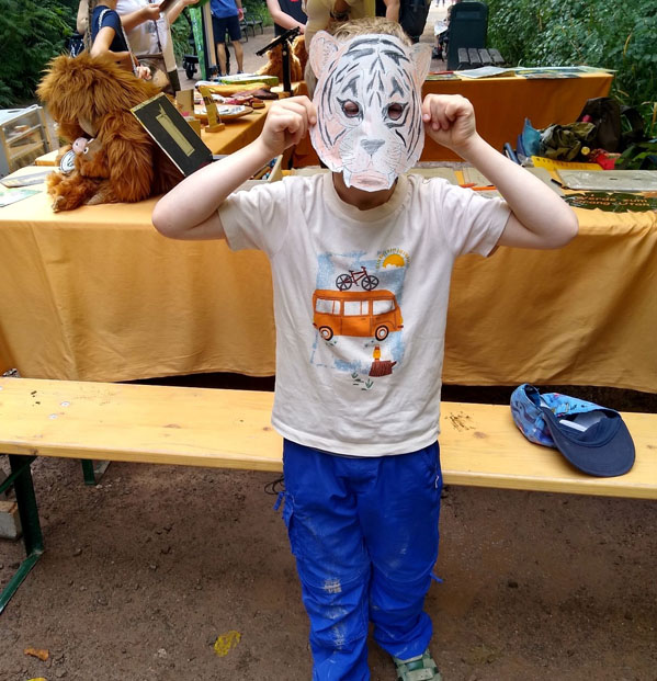 Vor dem Informationsstand des Orang-Utan in Not e.V. steht ein Kind und hält sich eine selbst gebastelte Tigermaske vor das Gesicht. Im Hintergrund unterhält sich ein Vereinsmitglied mit zwei Besucherinnen.