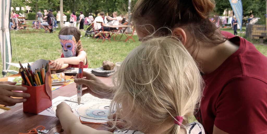 Eine Frau und zwei kleine Kinder sitzen an einem Tisch auf einer Wise und Malen mit Buntstiften. Eins der Kinder trägt eine bunte Orang-Utan-Maske. Im Hintergrund sitzen weitere Menschen an Tischen.