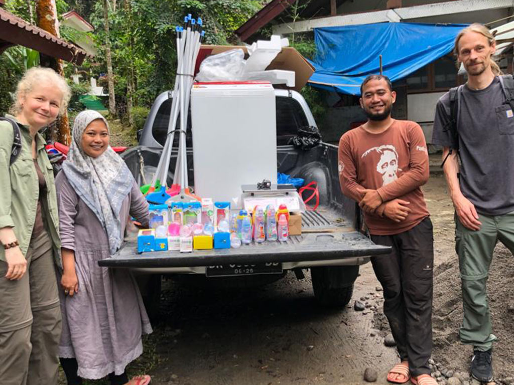 Übergabe von Nuckelfläschchen, einer Waschmaschine und weiteren Spenden. Zwei Vereinsmitglieder und zwei indonesische Helfer blicken freundlich in die Kamera.