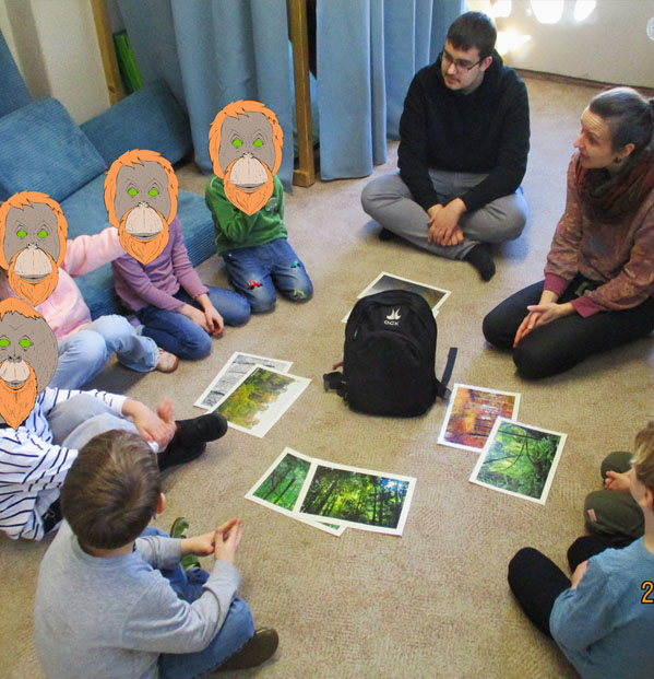 Mehrere Kinder und zwei Erwachsene sitzen im Kreis auf einem Teppich. In der Mitte befinden sich ein schwarzer Rucksack und Bildkarten von verschiedenen Jahreszeiten.