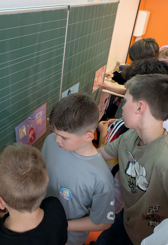 Mehrere Kinder stehen an einer Tafel und bekleben Bildkarten mit Klebepunkten. Auf den Bildkarten sind Geschenke, Bastelpapier, Toilletenpapier und weitere Dinge aus Papier abgebildet.
