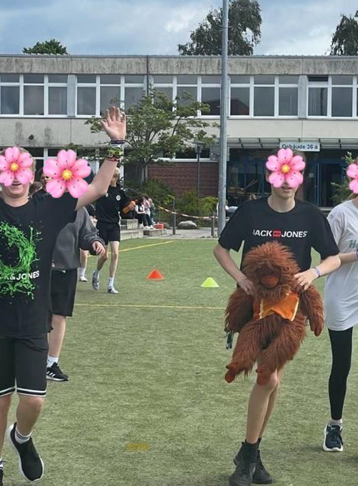 Eine Gruppe Schülerinnen und Schüler laufen auf einem Sportplatz. Ein Junge trägt eine Orang-Utan-Handpuppe.
