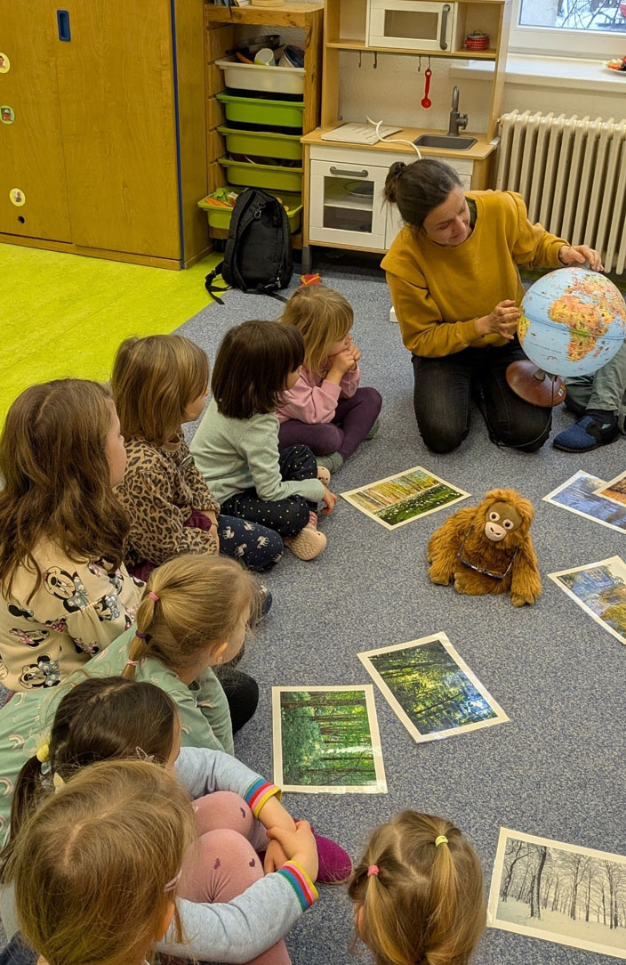 Kinder sitzen auf dem Boden. Darauf liegen Bildkarten von Jahreszeiten und ein Orang-Utan-Plüschtier. Die Umweltbildnerin zeigt etwas auf einem Globus.